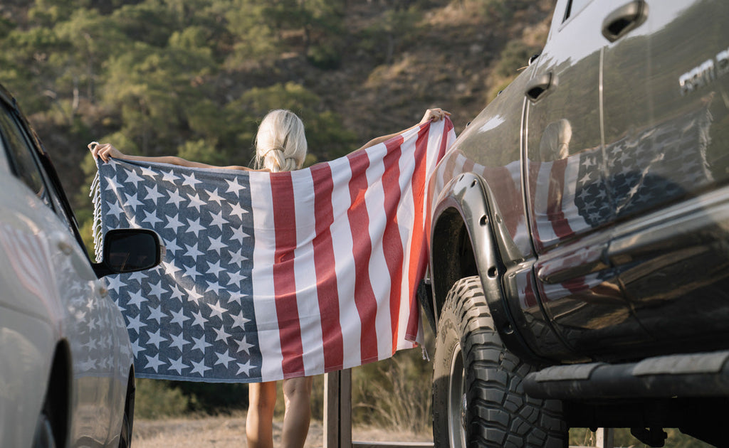 Person holding an American flag beach towel between two parked trucks – Wholesale Beach Towels USA