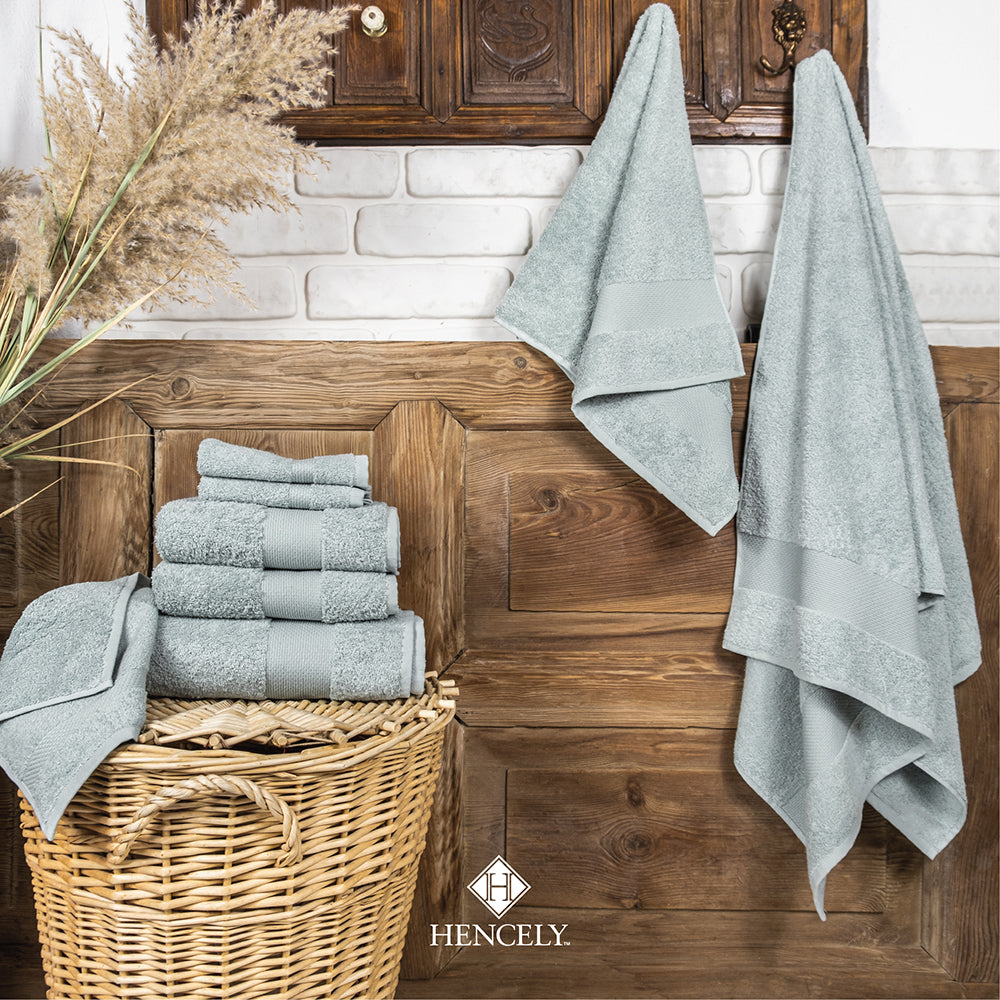 Light green Turkish cotton bath sheets displayed in a rustic bathroom setting, with folded towels stacked in a wicker basket and additional pieces hanging on wooden hooks against a white brick wall.
