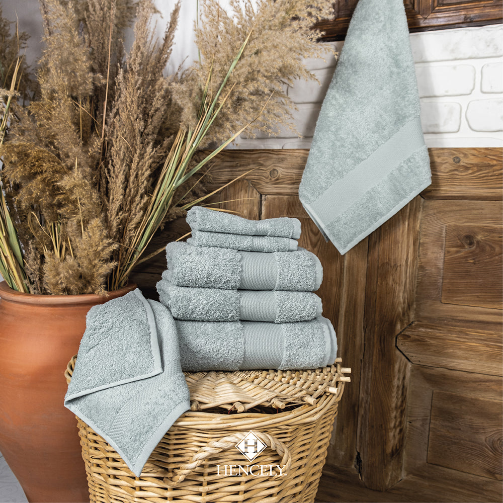 Stack of light green Turkish cotton bath sheets neatly folded in a wicker basket, with one towel hanging on a wooden wall and decorative dried plants adding a rustic bathroom style.