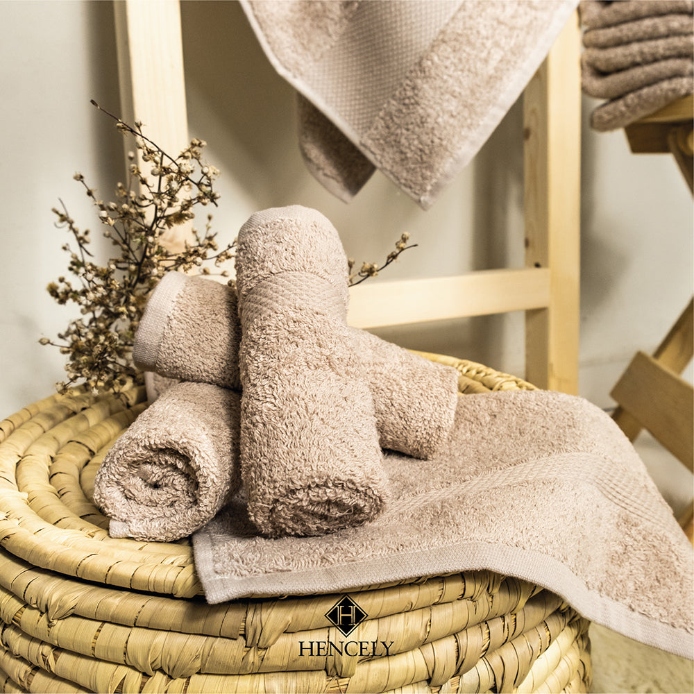 Rolled beige Turkish cotton bath sheets arranged in a woven basket with decorative dried flowers, displayed in a cozy bathroom setting by Hencely.