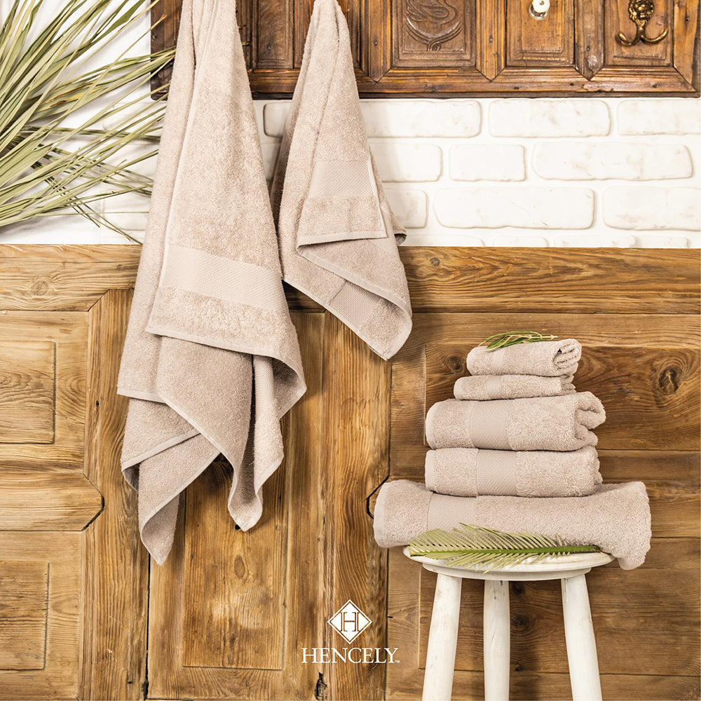 Hencely beige Turkish cotton bath sheets stacked on a stool and hanging on rustic wooden wall for bathroom decor.