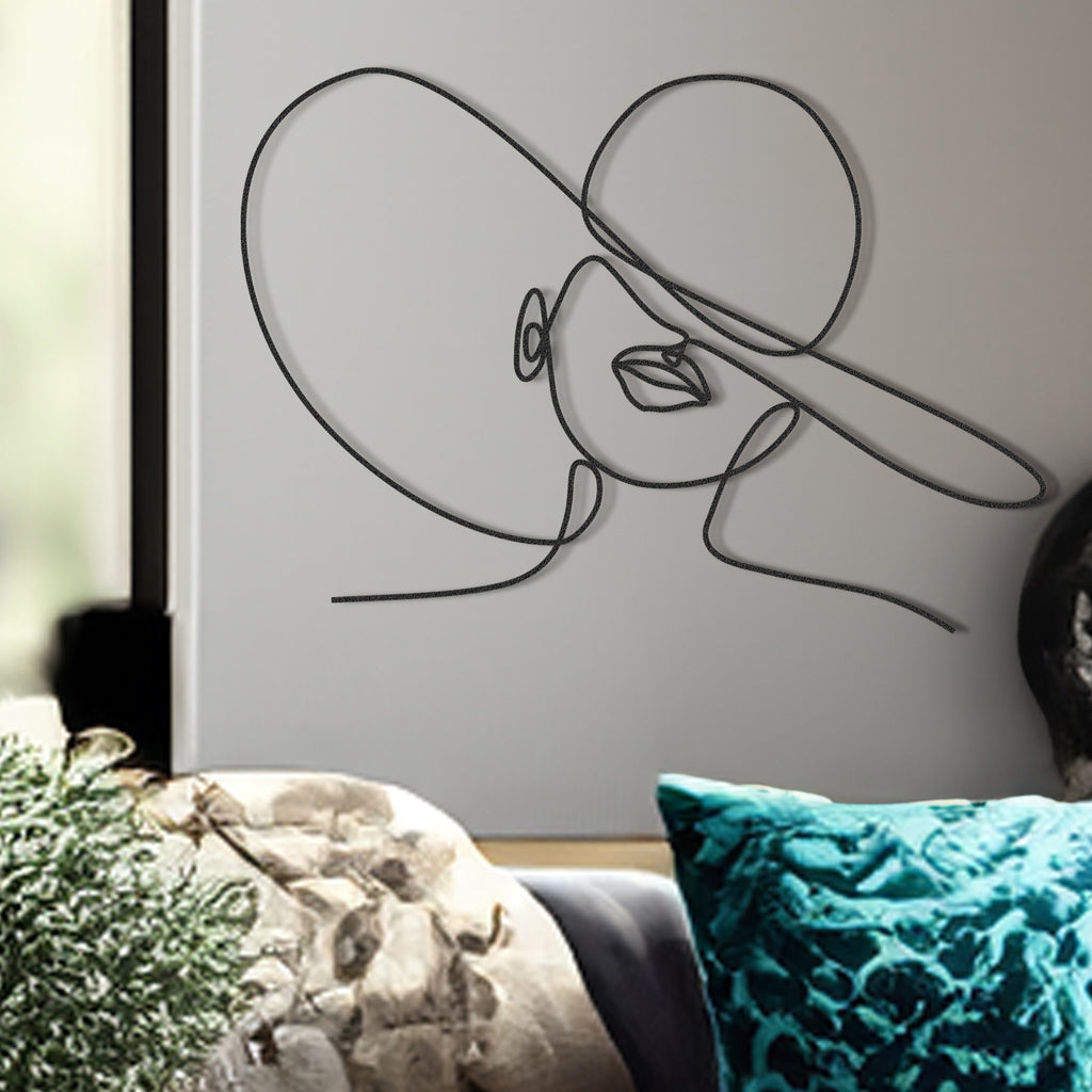 A minimalist metal wall art depicting a one-line drawing of a woman's face with a hat, in matte black color, displayed on a wall above a sofa with cushions.