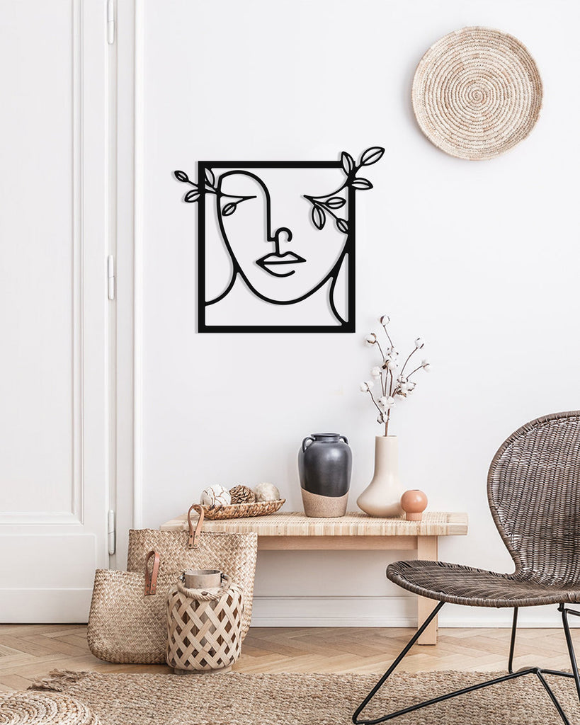 Black line art metal wall decor of a female silhouette with flowers, displayed on a white wall above a wooden bench with vases, baskets, and a woven chair.