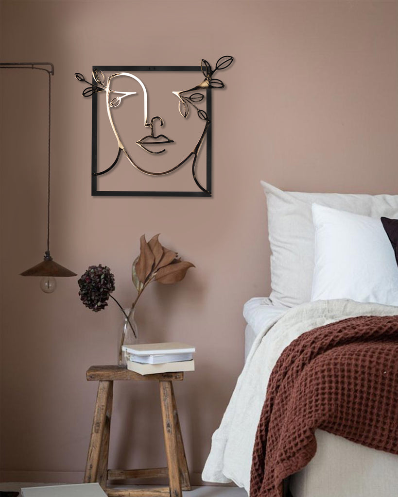 Gold line art metal wall decor of a female silhouette in a wide-brimmed hat, displayed on a soft beige wall above a bedside table with flowers and books.
