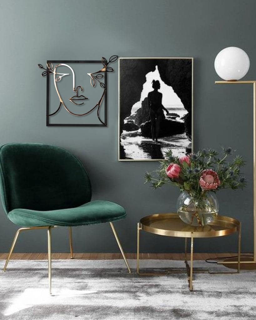 A gold line art metal wall decor of a female silhouette in a wide-brimmed hat, displayed on a dark wall above a green chair and a gold side table, with a black and white image in the background and a vase of flowers on the table.
